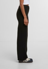 Černé kalhoty s vysokým pasem a širokými nohavicemi z měkkého pleteného materiálu, s žebrovaným páskem a hladkým povrchem. Nosí se s pohodlnou obuví.