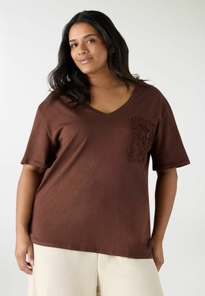 Donna con lunghi capelli scuri che indossa una maglietta marrone con scollo a V e una tasca testurizzata, abbinata a pantaloni crema, su uno sfondo semplice.