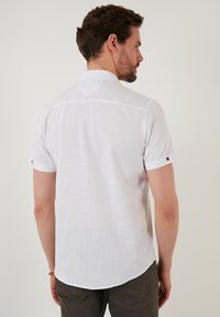Camisa blanca de manga corta hecha de tela texturizada, con cuello, mangas enrolladas con detalles de botones y un dobladillo ligeramente curvado en la parte posterior.