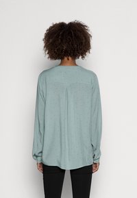 Blouse bleu clair à manches longues, avec un col rond et de subtils pois noirs. Tissu lisse avec une coupe décontractée.