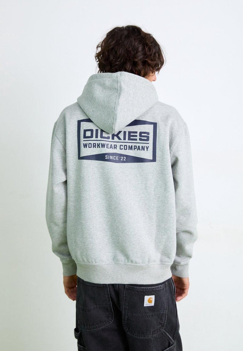 Personne portant un sweat à capuche gris clair avec l'inscription « Dickies Workwear Company Since '22 » en texte bleu marine dans le dos, associée à un pantalon de travail noir.
