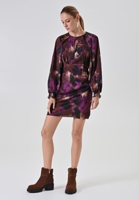 Lila- und braun gemustertes Kleid mit rundem Ausschnitt und Ballonärmeln. Kombiniert mit braunen Wildleder-Stiefeletten mit seitlichem Reißverschluss.