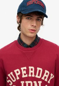 Czerwona bluza z dużym kremowym napisem "SUPERDRY", zestawiona z kołnierzykiem w kratę i niebieską czapką z tym samym logo marki.