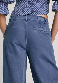 Pantalones de pierna ancha en azul claro con textura suave, que cuentan con un bolsillo trasero y detalle de costura, confeccionados en una tela resistente.