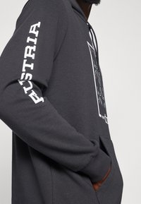 Felpa grigia scura con cappuccio, caratterizzata da una tasca frontale, polsini elastici e una stampa bianca "AUSTRIA" sulla manica sinistra. Design grafico sul davanti.