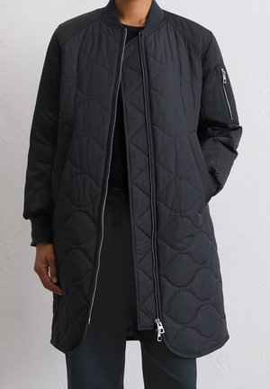 Veste bomber longue noire matelassée avec fermeture éclair frontale et poche zippée sur la manche, portée sur un pantalon et un haut noirs.
