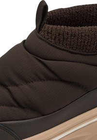 Gros plan d'une chaussure d'hiver isolée marron foncé avec tissu rembourré, languette texturée et col doublé en polaire au-dessus d'une semelle beige.