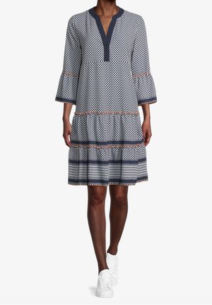 Kobieta w granatowo-białej sukience z wzorem sięgającej do kolan, z rozszerzanymi rękawami i białych trampkach, idąca do przodu na białym tle.