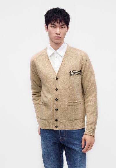 Jeune homme portant un cardigan beige boutonné avec un logo écusson sur une chemise blanche et un jean bleu, debout devant un fond blanc uni.