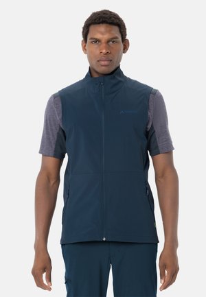 Uomo che indossa un gilet senza maniche blu navy con cerniera sopra una maglietta grigia a maniche corte, in piedi davanti a uno sfondo bianco uniforme.