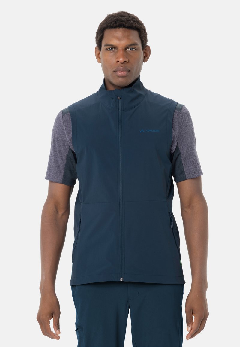 Mann trägt marineblaues ärmelloses Reißverschluss-Weste über einem kurzärmligen grauen Hemd und steht vor einem einfarbigen weißen Hintergrund.