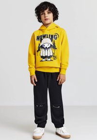 Felpa gialla con stampa grafica che presenta un design di un orso, abbinata a pantaloni jogger neri con aperture alle ginocchia e scarpe da ginnastica bianche.