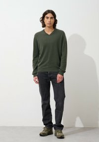 Groene v-hals trui van een zachte stof, gecombineerd met een donkere spijkerbroek en olijfgroene sneakers. Model staat tegen een effen witte achtergrond.