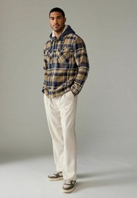 Geruite, beige en marineblauwe flanellen shirt met een grijze capuchon, gecombineerd met witte broeken en bruine sneakers met witte accenten.