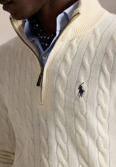 Uomo che indossa un maglione crema lavorato a trecce con cerniera, una camicia blu con il colletto e una sciarpa a pois sotto.