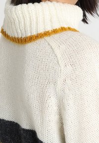 Gestrickter Pullover mit hohem, geripptem Kragen in Creme mit Senffarbener Einfassung. Der Körper zeigt horizontale Streifen in Creme, Grau und Schwarz.