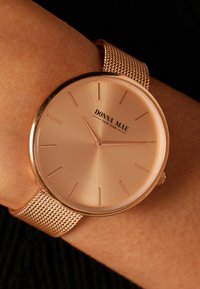 Ronde horloge met een metallic wijzerplaat in roségoud, minimalistische uuraanduidingen en een gestructureerde roségouden meshband om een pols.