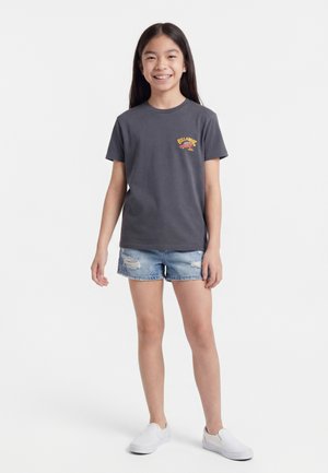 Fiatal lány mosolyog, sötétszürke Billabong pólót visel, világoskék szakadt farmer rövidnadrágot és fehér bebújós cipőt, fehér háttér előtt áll.