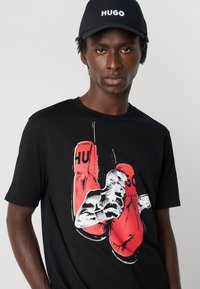Bărbat purtând o șapcă neagră cu textul "HUGO" și un tricou negru cu un grafic care prezintă mănuși de box roșii și un boxer în acțiune.