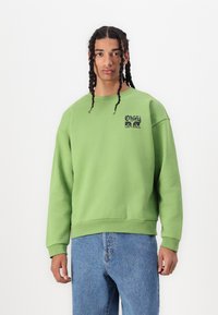 Hanorac verde cu manșete și tiv din elastan, având un logo grafic negru pe piept. Asortat cu blugi din denim de culoare albastru deschis.