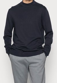 Maglione girocollo blu navy in tessuto morbido con polsini e orlo a costine, abbinato a pantaloni grigio chiaro con vestibilità sartoriale.