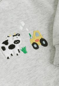 Bestickte schwarz-weißes Kuh neben grünem Gras und einem gelben Traktor auf hellgrauem Stoff.
