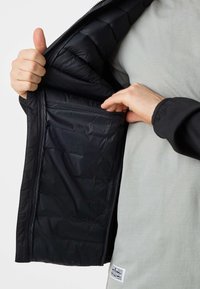 Hand opening black jacket to show inside chest pocket, wearing light gray shirt with a small white label near the waist.