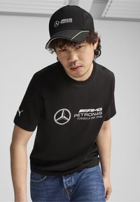 Juodas medvilninis marškinėliai su baltu AMG Petronas Formula One logotipu, derinami su juoda kepure su žaliais akcentais ir logotipu priekyje.