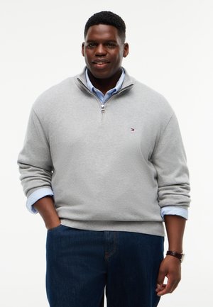 Mand med mørk hud iført en lysegrå sweater med kvart-lynlås over en blå skjorte og mørke jeans, med den ene hånd i lommen og den anden ved siden.