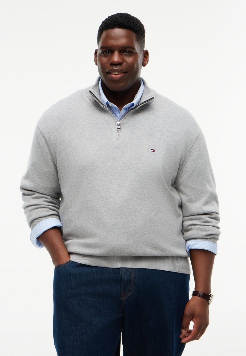 Homme à la peau foncée portant un pull gris clair à fermeture éclair quart, par-dessus une chemise bleue et un jean foncé, une main dans la poche et l’autre le long du corps.