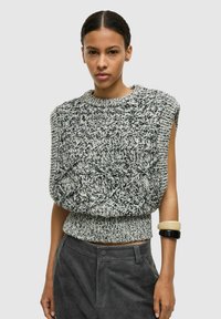 Strikket ærmeløs sweater med et tekstureret sort-hvidt mønster, rund halsudskæring og ribbet kant. Modellen bærer stabler af armbånd.