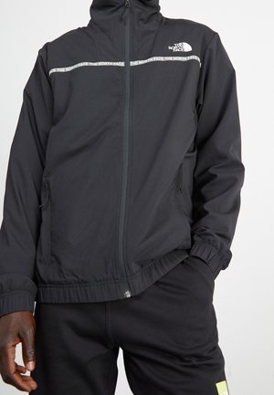 Sort lynlåstrøje med "The North Face" logo og gentagen branding over brystet, båret med sorte bukser og en hånd i lommen.