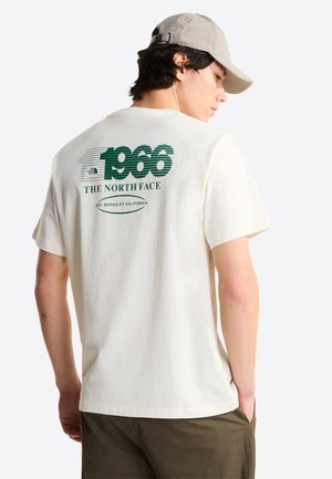 Nuori mies, joka pukeutunut valkoiseen t-paitaan, jossa on takana teksti "1966 The North Face Est. Berkeley California", yhdistettynä beigen väriseen lippalakkiin ja vihreisiin housuihin.