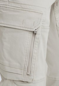 Beige cargo pants with a zippered pocket, stitched details, and a soft texture, featuring multiple spacious pockets for utility.