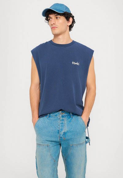 Tricou fără mâneci de culoare bleumarin cu logo alb, asortat cu o pereche de pantaloni din denim de culoare bleu deschis, cu buzunare și șnur. O șapcă albastră finalizează ținuta.