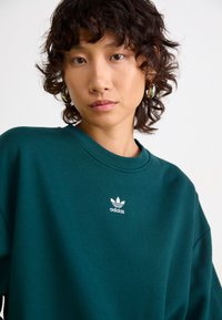Jeune personne ayant les cheveux courts et bouclés, portant des boucles d'oreilles en argent et un sweat à capuche Adidas vert foncé avec le logo au centre de la poitrine.