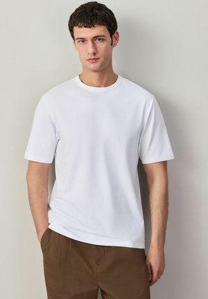 Mladý muž s krátkymi tmavými vlasmi, oblečený v obyčajnom bielom tričku s krátkym rukávom a hnedých nohaviciach, stojaci pred svetlosivou stenou.