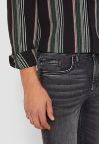 Črna majica s črnimi in rdečimi navpičnimi črtami na zavihku rokava, v kombinaciji s temno sivo denim hlačami, ki imajo subtilno obrabo in oznako logotipa.