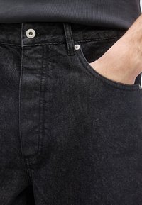 Χέρι στην τσέπη μαύρου τζιν με εμφανές κουμπί, καρφιά και θηλιά ζώνης, φοράει σκουρόχρωμο πλεκτό πουκάμισο μέσα από το παντελόνι.