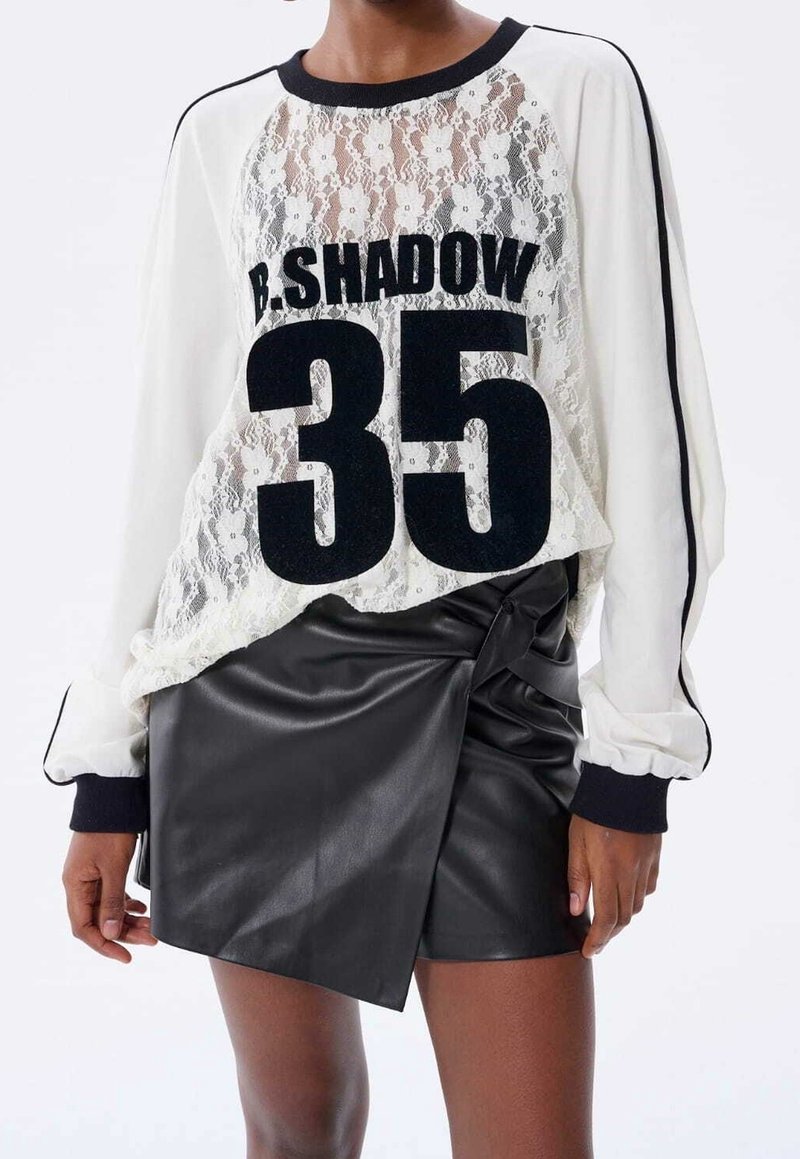 Personne portant un haut blanc en dentelle à manches longues avec un texte noir "B. SHADOW 35" et une jupe portefeuille noire en similicuir.