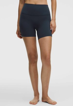 Femme portant un short de sport noir ajusté taille haute, debout pieds nus sur un fond uni gris clair.
