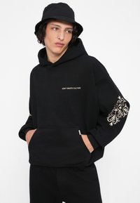 Melns hoodie ar uzrakstu "NEIZTĪDI KULTŪRU" uz krūšu daļas un greznu krēma krāsas rakstu uz piedurknes. Kangalas kabata, mīksta auduma, brīva izgriezuma.