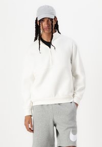 Felpa bianca con zip e colletto alto, abbinata a pantaloni corti grigi con un logo bianco. Tessuto liscio e vestibilità rilassata. Cappellino grigio indossato.