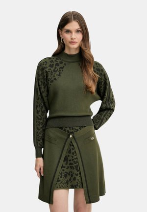 Donna che indossa un maglione verde oliva e una gonna coordinata con stampa leopardata nera e un flap anteriore decorativo con chiusura a bottone.
