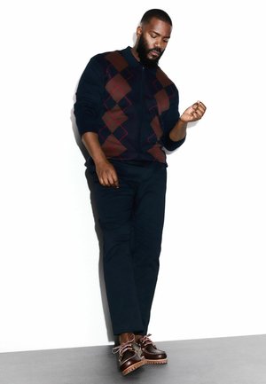 Laivaston sininen vetoketjupaita, jossa on timanttikuosi burgundin ja ruskean sävyissä, yhdistettynä tummiin housuihin ja ruskeisiin nauhallisiin kenkiin, joissa on teksturoidut pohjat.