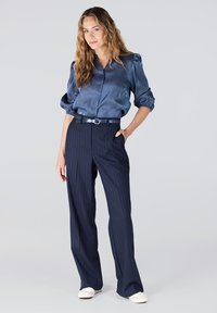 Navy satijnen blouse met knoopsluiting en pofmouwen, gecombineerd met navy streepbroek en witte sneakers, met een strakke riem.
