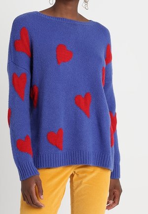 Pull en laine bleu avec des motifs de cœurs rouges porté par une personne, assorti à un pantalon en velours côtelé jaune moutarde.