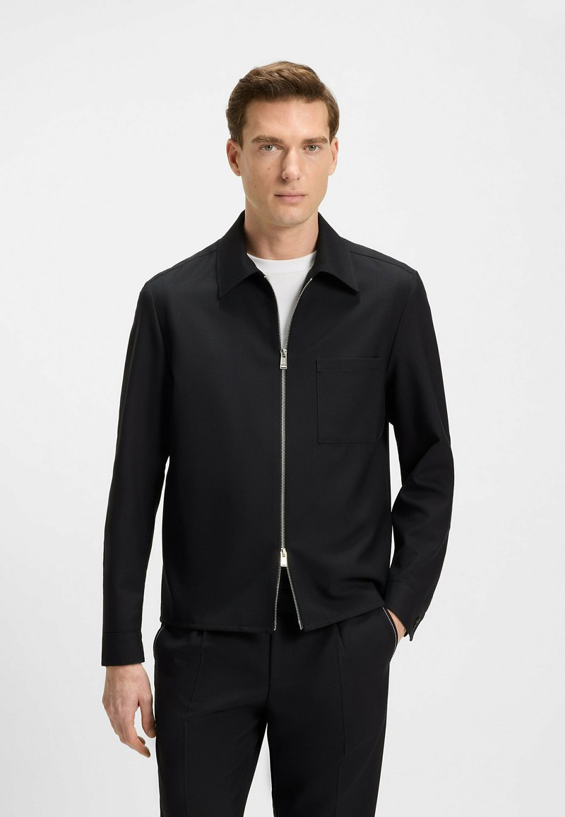 Black zip-up jacket made of smooth fabric. Features a collared neckline, single chest pocket, and minimalistic design. Clean lines and tailored fit.