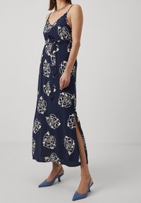 Kvinna i en marinblå blommig midiklänning med midjeband och sidofrans, kombinerad med blå slingback-klackar och ett guldhalsband.