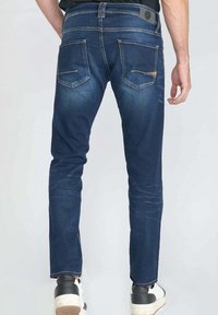 Jean en denim bleu foncé à coupe slim, avec poches arrière et détails de décoloration légère. Porté avec des baskets blanches et noires.